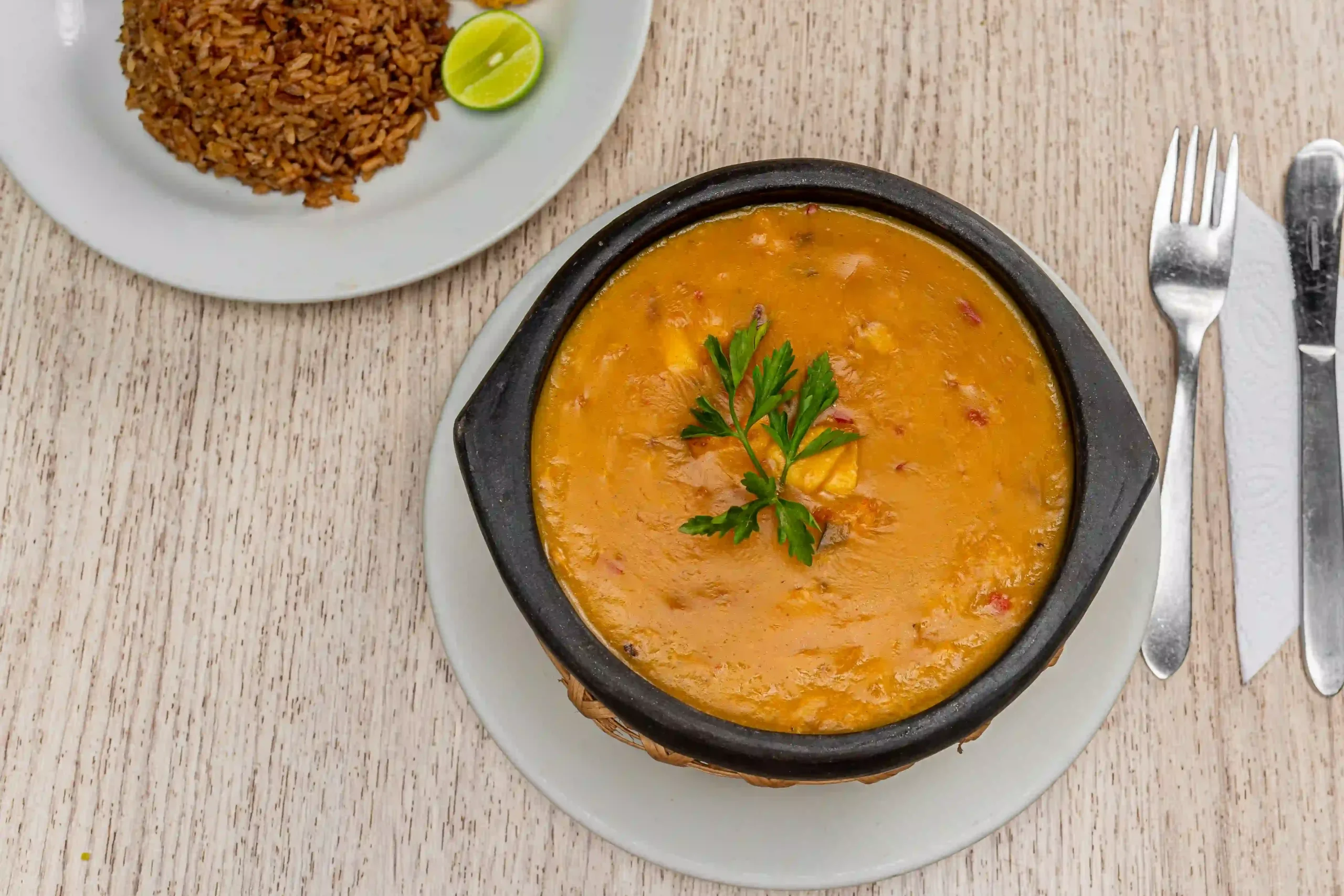 Sopa de mariscos servida en una cazuela de barro, acompañada de arroz de coco, patacones y rodajas de limón en un restaurante.