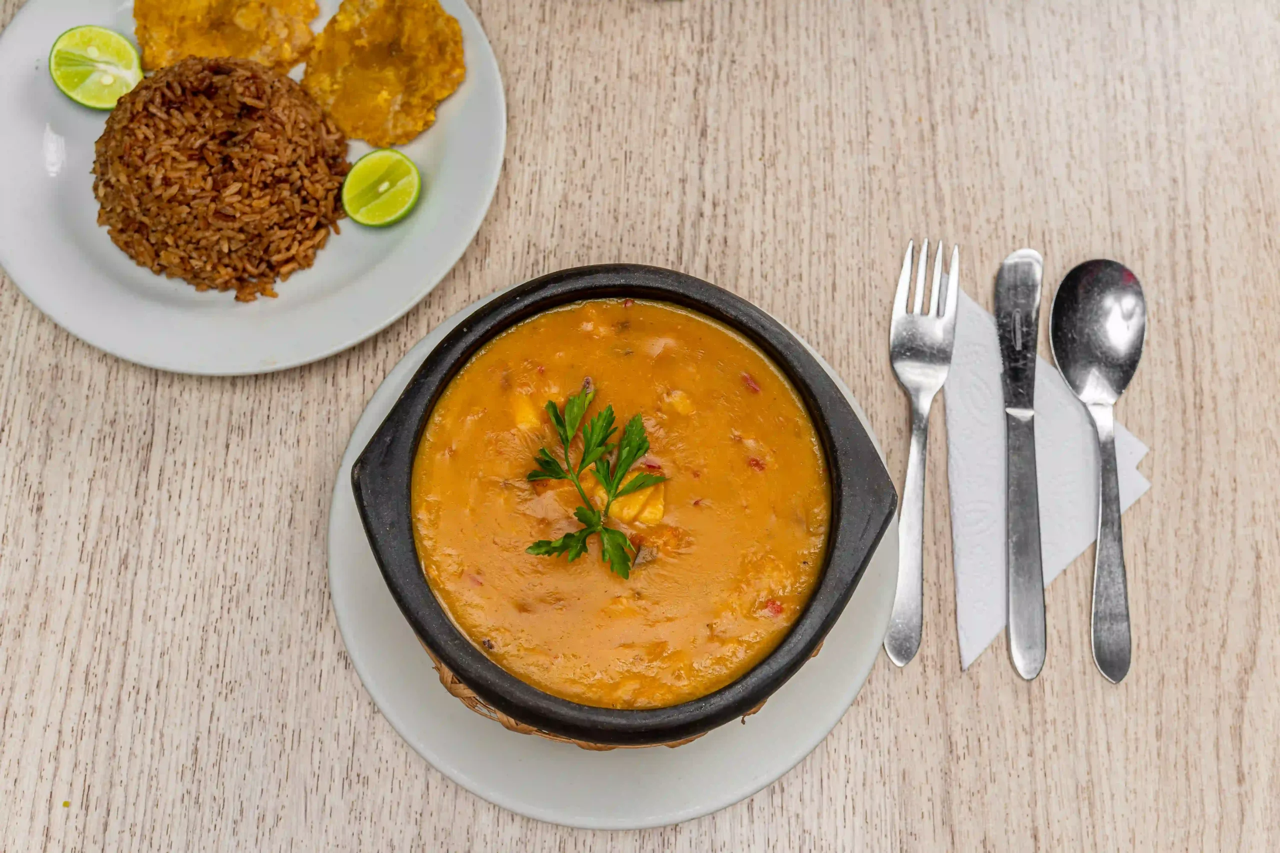 Sopa de mariscos servida en una cazuela de barro, acompañada de arroz de coco, patacones y rodajas de limón en un restaurante.