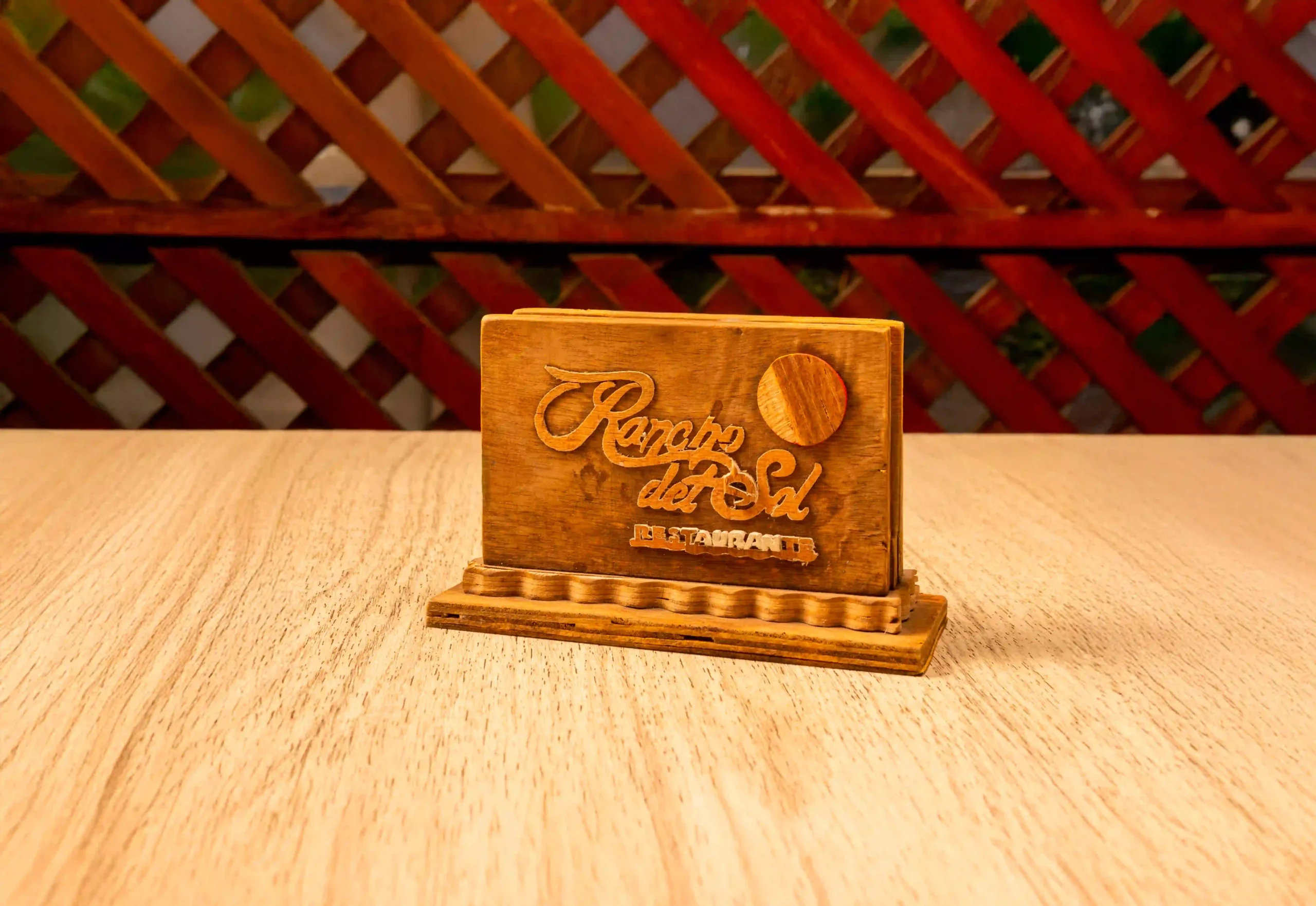 Porta servilletas de madera con el nombre y logo del restaurante "Rancho del Sol", colocado sobre una mesa de madera en un entorno rústico.