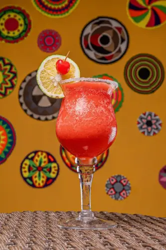 Cóctel frozen rojo decorado con rodaja de limón y cereza, servido en copa alta sobre mesa de mimbre con fondo de motivos artesanales.