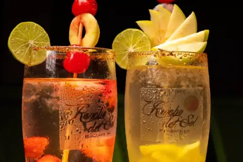 Dos cocteles frutales servidos en vasos de Rancho del Sol Restaurante Bar, decorados con frutas tropicales como lima, cereza, manzana y durazno.