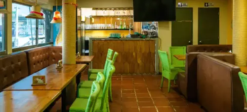 Interior de un restaurante con sillas tejidas verdes, mesas de madera, lámparas decorativas y un bar con estantería de cristalería y bebidas.